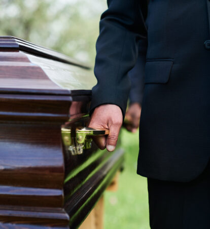 Close-up of man in black suit holding by handle of wooden coffin with dead person while carrying it with other people at funeral ceremony